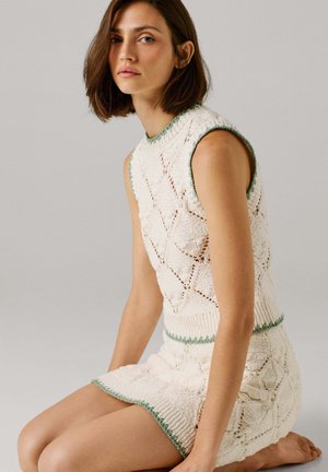 Frau mit kurzen braunen Haaren, die ein ärmelloses weißes gestricktes Oberteil und einen passenden Rock mit grünem Rand trägt, sitzt barfuß auf einem hellgrauen Boden.