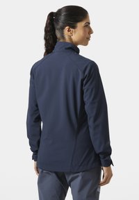 Kobieta o ciemnych włosach związanych w kucyk, ubrana w granatową kurtkę i szare spodnie, stoi tyłem i patrzy w prawo na białym tle.