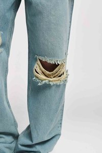 Heledad sinine teksapüksid, millel on suur lahtine horisontaalne rebend, mis paljastab naha põlve juures, heleda tausta ees.