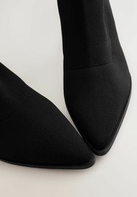 Juodi megzti kulkšniniai batai su smailiu pirštu ir tekstūruota paviršiumi. Turi tvirtą juodą padu su minimaliu akcentų detalėmis.