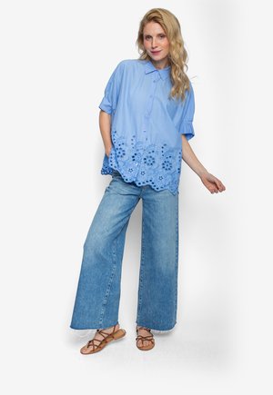 Frau trägt eine hellblaue Kurzarmbluse mit Blumen-Ausschnitten, weite Jeans und braune Riemchensandalen, steht vor weißem Hintergrund.