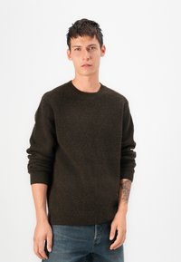 Suéter verde-escuro canelado com decote redondo e mangas longas, combinado com jeans azuis. O tecido parece suave e com um acabamento texturizado.