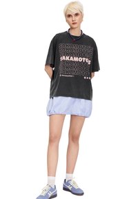 Černé oversized tričko s bílým a růžovým textem, doplněné světle modrou proužkovanou sukní. Model má na sobě modré tenisky s bílými akcenty.