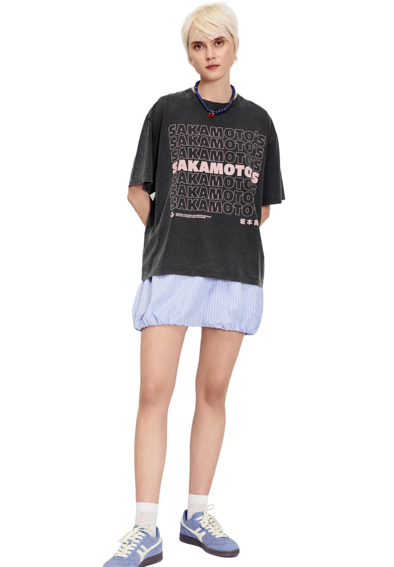 Černé oversized tričko s bílým a růžovým textem, doplněné světle modrou proužkovanou sukní. Model má na sobě modré tenisky s bílými akcenty.