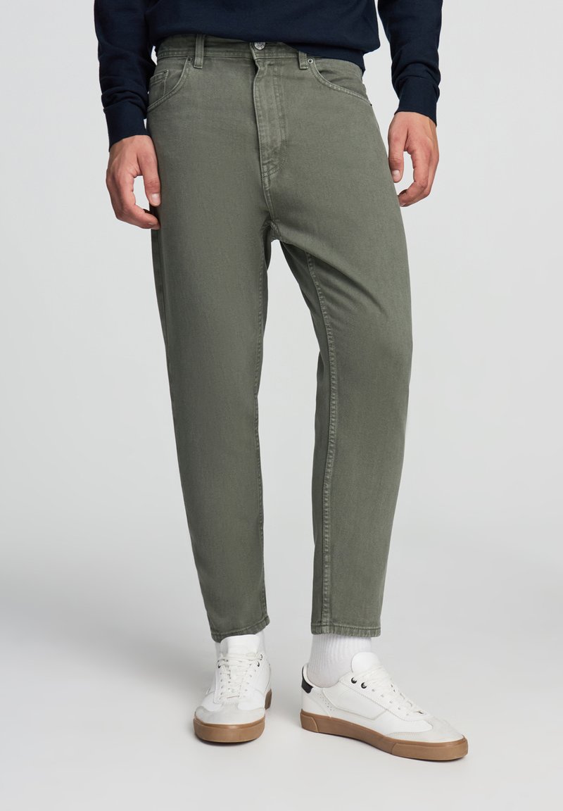 Pantaloni verde oliva in denim, con vestibilità dritta, cinque tasche e chiusura con bottone, abbinati a sneaker bianche.