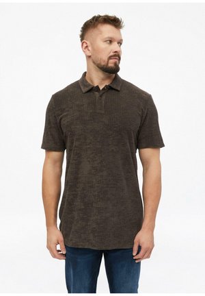 Mann mit kurzen Haaren und Bart, der ein dunkelbraunes kurzärmeliges Poloshirt und blaue Jeans trägt, schaut nach links vor weißem Hintergrund.