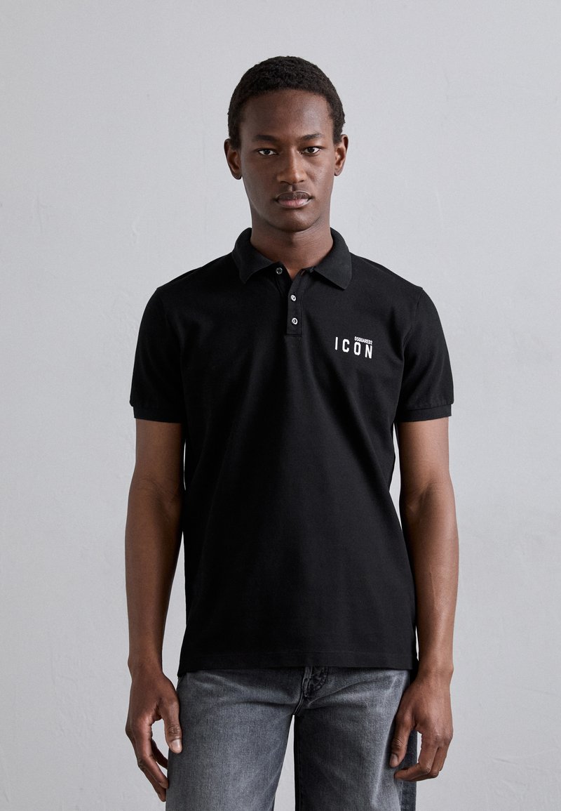 Schwarzes Poloshirt mit Kragen, das weißen "ICON"-Text auf der linken Brustseite trägt. Kombiniert mit grauen Jeans.