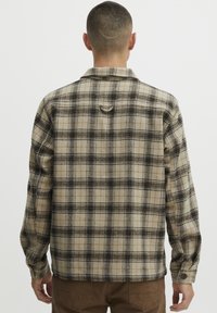 Chemise à carreaux avec une base beige, des motifs à carreaux noirs et bruns, des poignets boutonnés et une boucle sur le dos. Matière en coton, coupe décontractée.