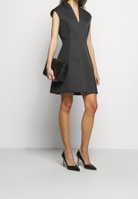 Kvinna som bär en ärmlös svart klänning med V-ringning, svarta spetsiga klackskor och håller en svart flätad kuvertväska, stående mot en vit bakgrund.