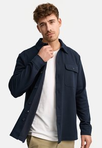 Chemise bouton montée bleu marine en tissu texturé, avec une poche poitrine et un col classique. Portée par-dessus un t-shirt à col rond blanc.
