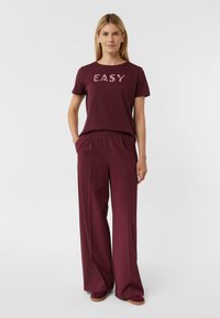 Maroon T-Shirt mit dem Text "EASY" in einem helleren Farbton. Kombiniert mit weit geschnittenen maroonfarbenen Hosen. Weicher Stoff mit bequemer Passform und elastischem Bund.