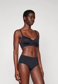 Femme avec les cheveux courts portant un soutien-gorge noir et un sous-vêtement noir assorti, se tenant contre un fond clair uni, faisant légèrement face à la gauche.