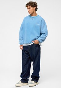 Lichtblauwe oversized sweatshirt met wit logo, gecombineerd met donkerblauwe baggy jeans en crèmekleurige sneakers. De outfit heeft een gelaagde styling.
