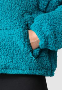 Petrolejový fleecový pulovr s texturovaným, měkkým povrchem. Má kapsu na zip a vytahovací brzdu pro nastavitelný střih.