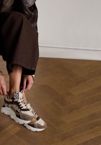 Klobige Sneaker mit braunen, beige und blauen Tierdruckakzenten, ausgestattet mit weißen Schnürsenkeln und einer dicken weißen Sohle, auf einem Holzboden.