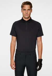 Svart pikétröja med krage och knappslå, korta raglan ärmar, ihop med svarta byxor och en svart handske på höger hand.