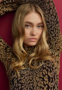 Top s leopardím vzorem v černé a zlaté barvě s vysokým výstřihem a dlouhými rukávy, vyrobený z měkké, vlnité textilie na burgundském pozadí.