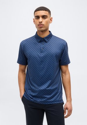 Mladi moški, oblečen v modro polo majico s kratkimi rokavi z vzorcem in črne hlače, stoji ob enobarvni svetli ozadju.