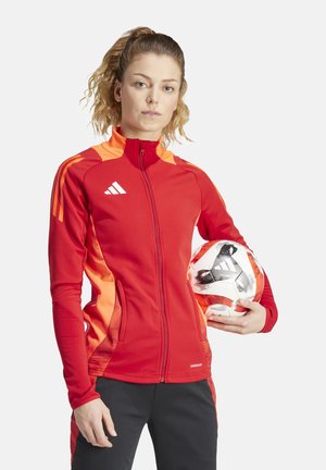 Giacca sportiva rossa con accenti arancioni, chiusura con zip, tasche con zip e branding AEROREADY, tenuta da una persona con un pallone da calcio.