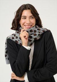 Sciarpa grigia con macchie leopardate nere e ruggine, realizzata in materiale morbido, drappeggiata attorno al collo. Indossata con un blazer nero e una camicia bianca.