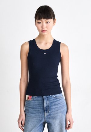 Jeune femme aux cheveux noirs et lisses, portant un débardeur bleu marine ajusté et un jean taille haute bleu clair sur fond blanc.