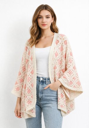 Frau trägt eine cremefarbene und rosa gehäkelte Cardigan mit offenem Vorderteil über einem weißen Tanktop und hellblauen High-Waist-Jeans, die Hand in der Tasche.