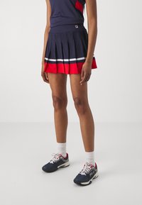 Mörkblå tennisplisserad kjol med röda och vita ränder vid fållen, kombinerad med en matchande ärmlös topp och mörkblå sportskor med grå detaljer.