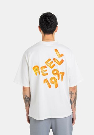 Man with short dark hair wears white oversized T-shirt with orange text "REELL 1997" on back, showing tattooed arms and light gray pants.