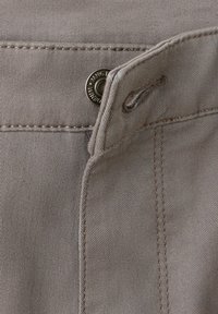 Gros plan sur un tissu en denim beige montrant un bouton métallique et une boutonnière sur une ceinture avec des coutures visibles.
