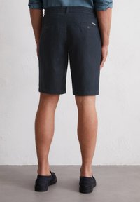 Navy blue linen shorts with a straight cut, two back pockets, and button closures. Worn with black slip-on shoes, displayed from the back.