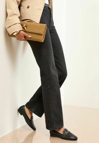 Schwarze Gerade-Jeans, beiger Trenchcoat, schwarze Loafers mit goldenen Beschlägen, hält eine hellbraune geflochtene Clutch vor einem hellen Hintergrund.