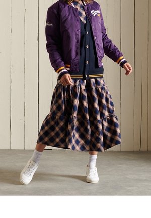 Giubbotto bomber viola con logo ricamato, cardigan blu navy, gonna a pieghe a strati in blu navy e beige, abbinata a sneaker bianche e calzini alla caviglia.