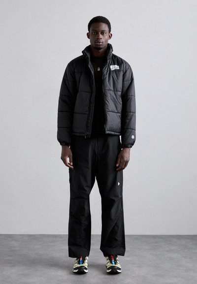 Svart puffjakke med kviltet design, glidelås, og logo; kombinert med løse svarte bukser og flerfargede joggesko. Glatt tekstur.