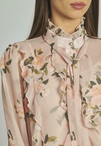 Blusa rosa chiaro con motivi floreali, bordi arricciati e colletto alto con un bottone decorativo al collo. Tessuto trasparente.