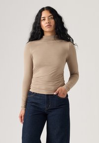 Beige, hosszú ujjú felső magas nyakkal, az deréknál összegyűrt, redőzött textúrával és karcsú, illeszkedő ujjakra, sötét farmernadrággal párosítva.