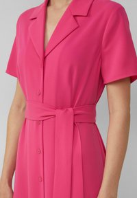 Roze jurk met korte mouwen, kraag en knoopsluiting. Heeft een ceintuur en is gemaakt van glad, lichtgewicht stof.