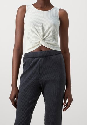 Vrouw draagt een mouwloos wit geribd cropped topje, voor gedraaid, gecombineerd met hooggetailleerde donkergrijze geribde broeken.