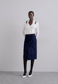 Witte lange mouwen top met uitsnijdingen, gecombineerd met een donkerdenim kokerrok met een split aan de voorkant. Het model draagt zwarte panty’s en witte hakken.