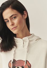 Svetlokrémová mikina s kapucňou so zapletenými sťahovacími šnúrkami a obrázkom medveďa na prednej strane. Má zapínanie na gombíky pri výstrihu.