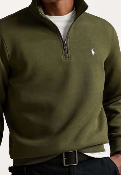 Olive green zippered pullover made of soft fabric, featuring a white logo on the left chest area and a layered white shirt underneath.