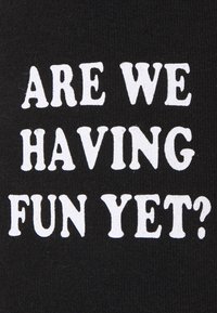 Černá látka s bílým textem "UŽ SE BAVÍME?" ve tučném, hravém písmu. Materiál vypadá měkce s hladkou texturou.