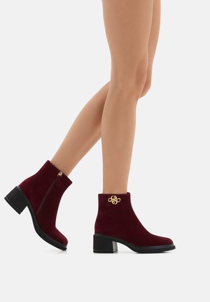 Beine, die burgunderfarbene Wildleder-Ankle Boots mit einem goldenen Dekorationsdetail und Blockabsätzen tragen, vor einem weißen Hintergrund gezeigt.