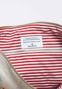 Interno della borsa di colore crema con rivestimento a righe rosso e bianco, dotato di un'etichetta che riporta "Fatto a mano in Germania" e una tasca con zip.