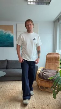 T-shirt branca de manga curta com um pequeno logótipo, combinada com jeans de perna larga azul-escuro e sapatos cinzentos de slip-on. Ambiente interior natural.