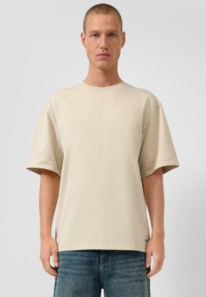 Mężczyzna z krótkimi włosami, ubrany w gładki, beżowy, oversize'owy T-shirt oraz niebieskie dżinsy, stojący na tle jednolitego jasnego tła.