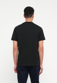 Άνδρας με κοντά σγουρά μαλλιά, φορώντας απλό μαύρο κοντομά�νικο T-shirt και σκούρους παντελόνες, εμφανίζεται από πίσω σε λευκό φόντο.