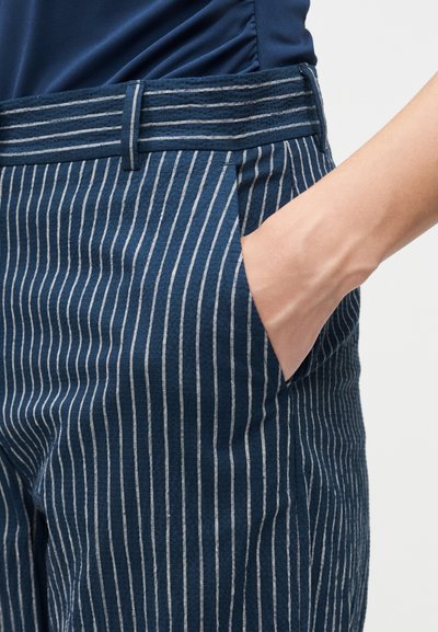 Χέρι που ακουμπά στην τσέπη μπλε ριγωτών παντελονιών με ταιριαστή ζώνη, φορεμένα με σκούρο μπλε πουκάμισο.