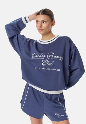Žena v modrej oversized mikine a rovnako ladených šortkách s textom „Cardio Bunny Club“, pózujúca s jednou rukou za hlavou.
