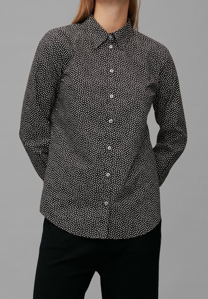 Femme portant une chemise noire à manches longues avec de petits pois blancs et un pantalon noir sur fond gris.