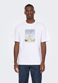 Λευκό βαμβακερό t-shirt με εκτυπωμένη εικόνα του Πύργου του Άιφελ σε απαλό, ξεθωριασμένο φόντο. Κοντά μανίκια και στρογγυλή λαιμόκοψη.